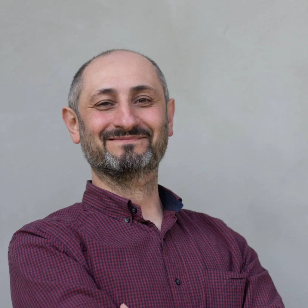 Wsparcie psychiczne dla młodzieży - Fundacja Hope. Mateusz Gwóźdź, członek zespołu Fundacji Hope. Uśmiechnięty mężczyzna w bordowej koszuli w kratę, z założonymi rękami, patrzący w obiektyw wzrokiem pełnym życzliwości i spokoju.