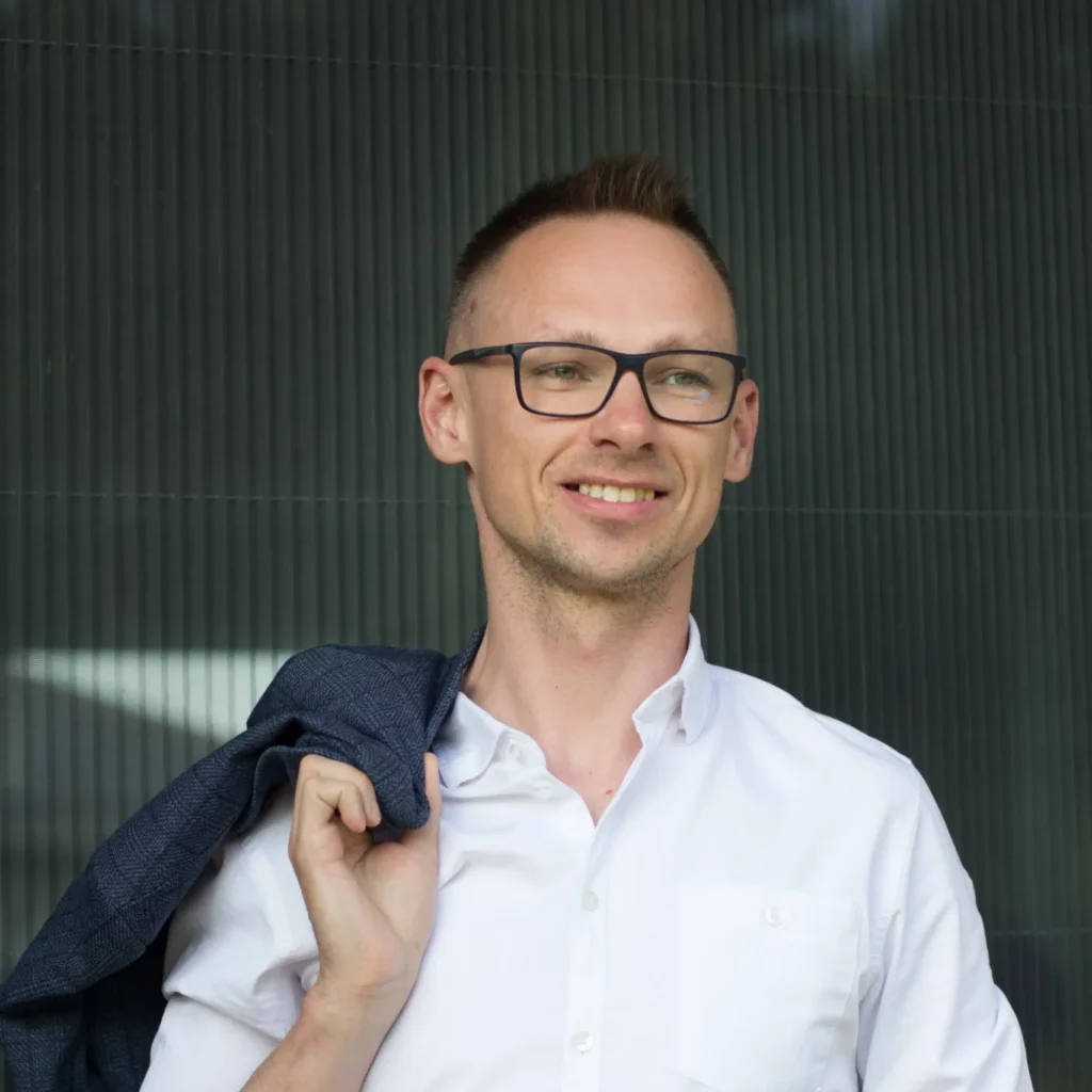 Wsparcie psychiczne dla młodzieży - Fundacja Hope. Tomasz Grzegorzek z Fundacji Hope. Mężczyzna w okularach i białej koszuli, trzymający marynarkę na ramieniu, uśmiecha się, patrząc w przyszłość z optymizmem.